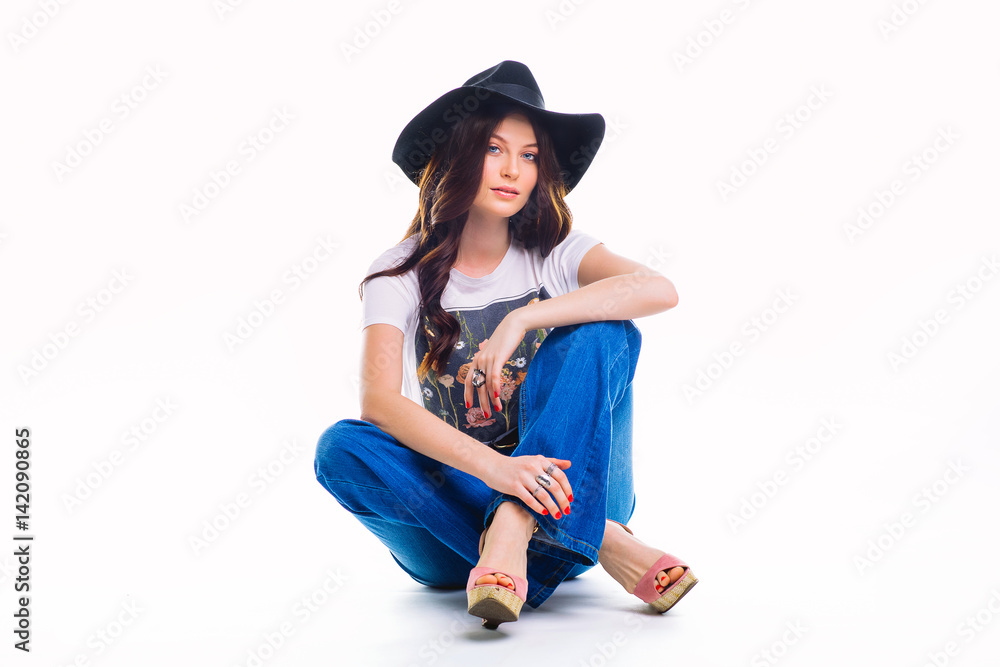Beautiful young brunette in jeans bell bottom white t-shirt and black hat ,sitting on floor in Studio isolated on white background. Beautiful young brunette in jeans bell bottom white t-shirt and black hat ,sitting on floor in Studio isolated on white background.
