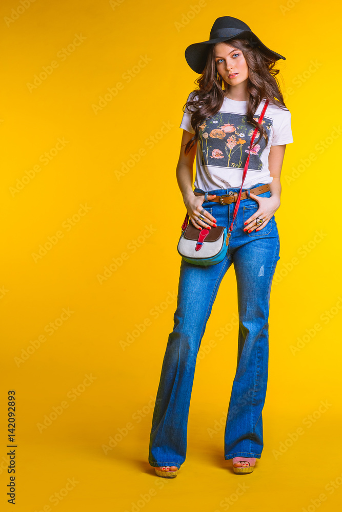 Beautiful young brunette girl in white t-shirt jeans flared with a small bag and a hat standing on a yellow background in the Studio.Fashion looks concept hats clothing elegance. Beautiful young brunette girl in white t-shirt jeans flared with a small bag and a hat standing on a yellow background in the Studio.Fashion looks concept hats clothing elegance.