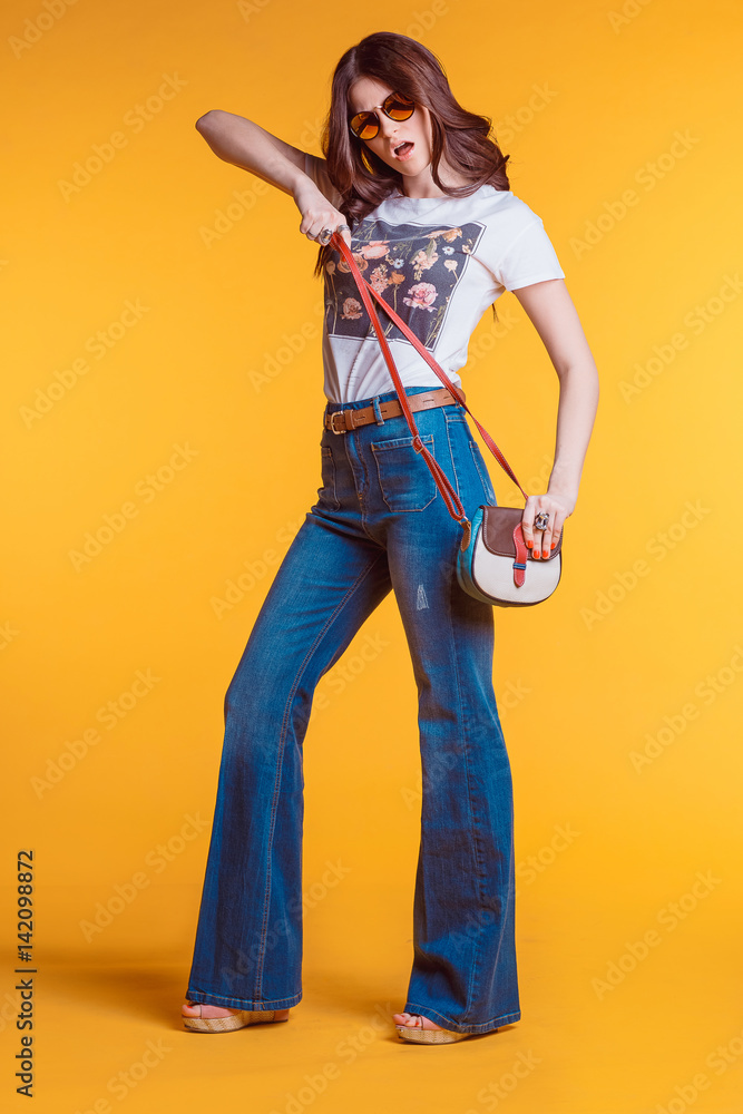 Beautiful young brunette girl wearing white t-shirt jeans flared with a small bag standing on a yellow background in the Studio..Bright stylish look Beautiful young brunette girl wearing white t-shirt jeans flared with a small bag standing on a yellow background in the Studio..Bright stylish look