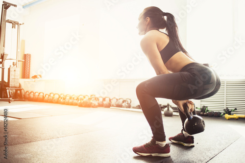 Athletic woman exercising with kettle bell while being in squat position. Muscular woman doing crossfit workout at gym.