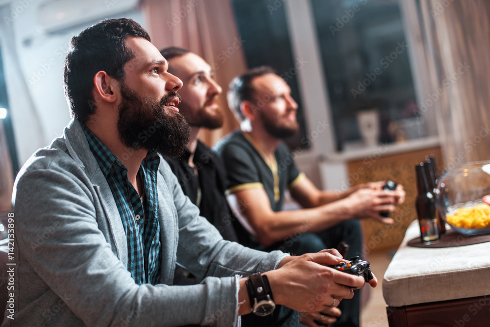 Men with a beard sitting on the couch at home with beer and chips with joysticks in hand playing computer video games. The concept of friendship, technology and weekend