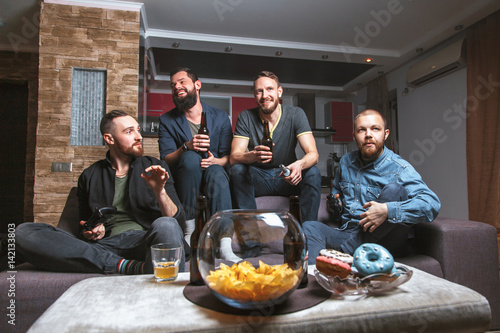 A company of four men with a beard sitting on the couch at home with beer and chips  two of them with joysticks in hand playing computer video games  the other carefully watching the game. 