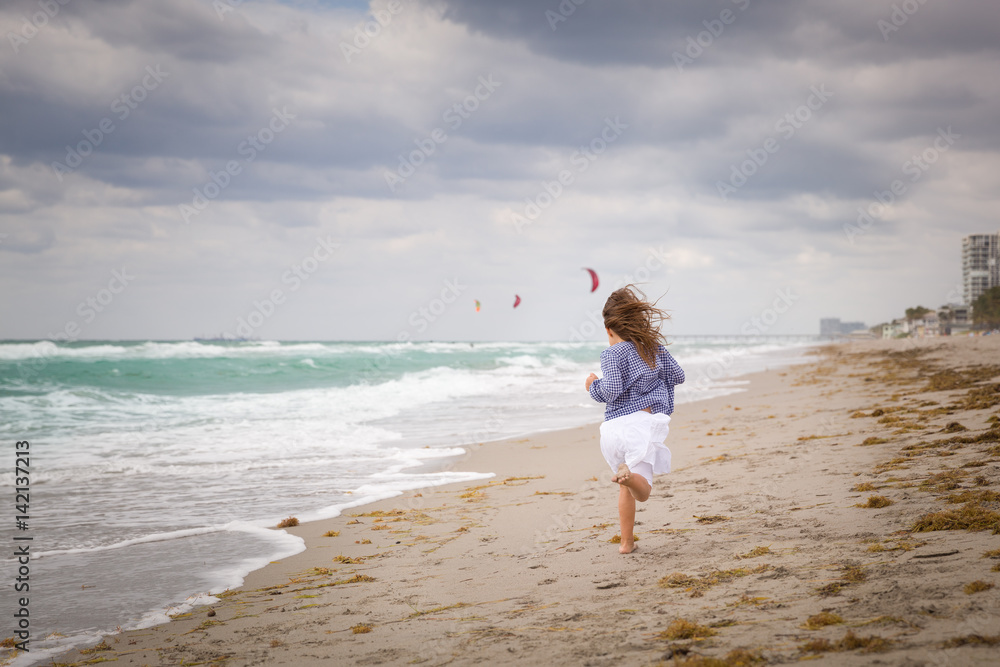 Little girl running on the ocean beach on a cloudy day. Vacation by the sea. Cute kid girl on the deserted beach. Summer, outdoors. Wind in the hair of a small girl. Beach landscape.