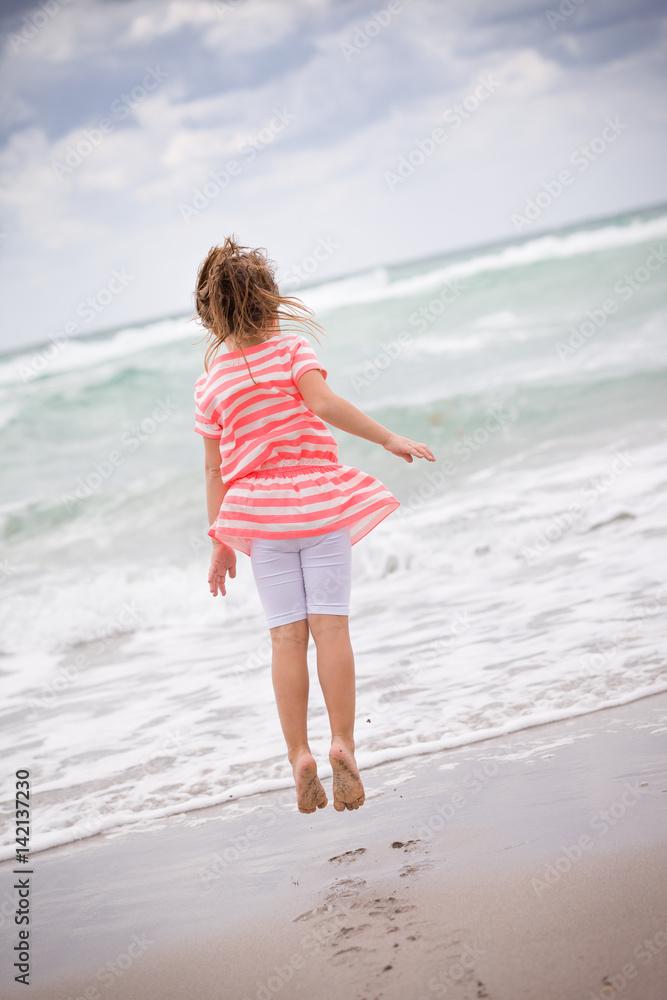 Little adorable kid girl in striped dress jumping on the sandy beach and having fun at the ocean. Sea vacations. Child dreaming. Outdoors. Back view.
