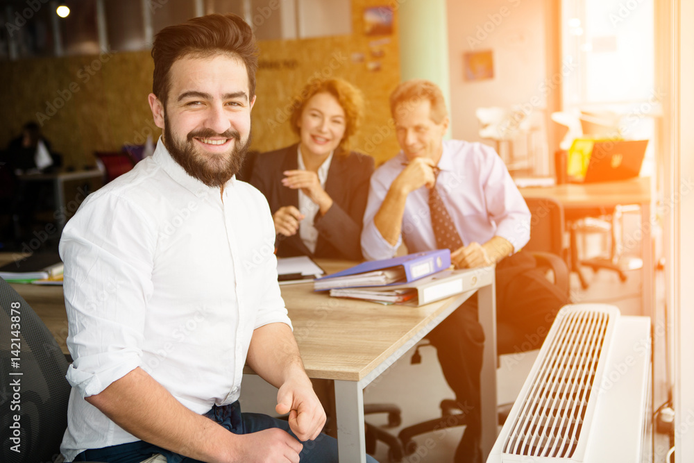 Happy entrepreneur man smiling after successful deal with huge company, firm, enterprise. His business colleagues sitting on background in office.