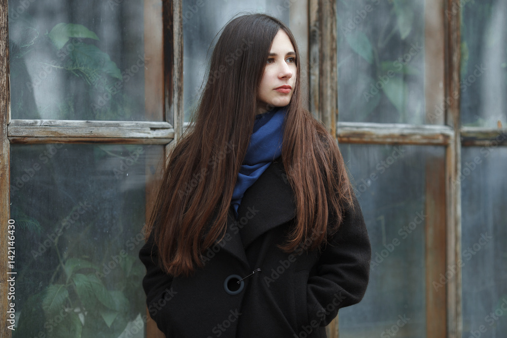 Fototapeta premium Young beautiful girl in a black coat and blue scarf for a posing in the autumn / spring park. An elegant brunette girl with gorgeous extra long hair. Lifestyle concept.