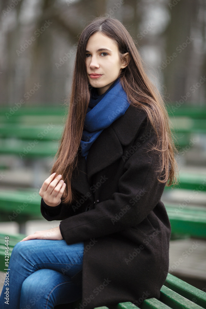 Young beautiful girl in a black coat blue scarf glasses sitting on bench in city park. An elegant brunette girl with gorgeous extra long hair. Vintage film effect saturated colors.