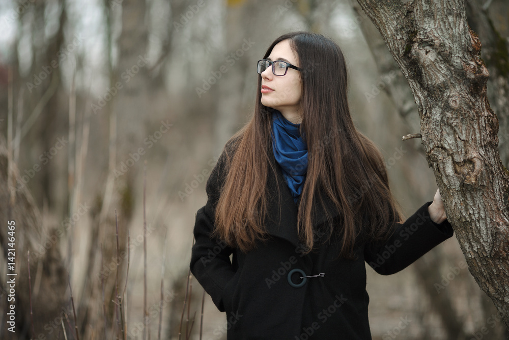 Young beautiful girl in a black coat blue scarf glasses walking in the  autumn / spring forest