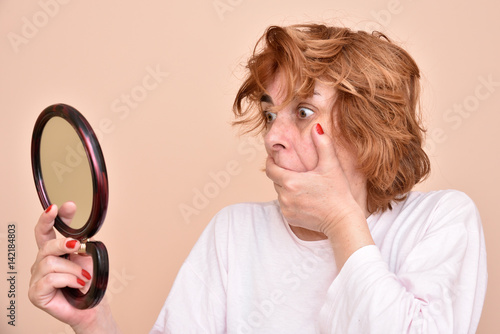 Shocked and surprised middle aged woman looking at her skin and unkempt hair in the mirror photo