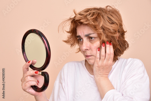 Unhappy and dissatisfied middle aged woman looking at her skin and unkempt hair in the mirror