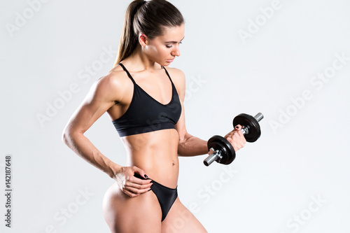Athletic woman in black clothing with muscular body with dumbbell over gray background.