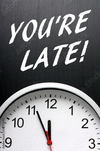 The words You're Late in white text on a blackboard above a wall clock as a reminder of the importance of being on time