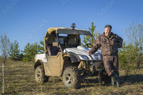 Male fisherman, hunter camouflage in a suit standing next to the ATV, SUV in forest, field, summer, men's Hobbies.