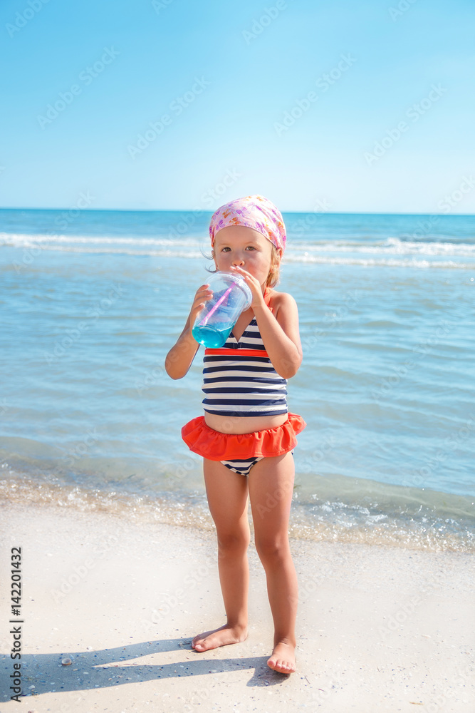 Happy little girl in swimsuit drink cocktail on the beach. Summer. Vacation. Sea. Ocean. Happy little girl in swimsuit drink cocktail on the beach. Summer. Vacation. Sea. Ocean.