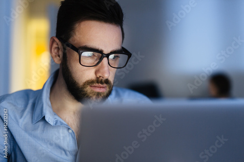 Bild auf Leinwand Portrait of handsome bearded man wearing glasses working with laptop in dark off