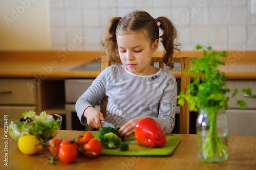 Cute girl of younger school age cuts vegetables and greens for salad.