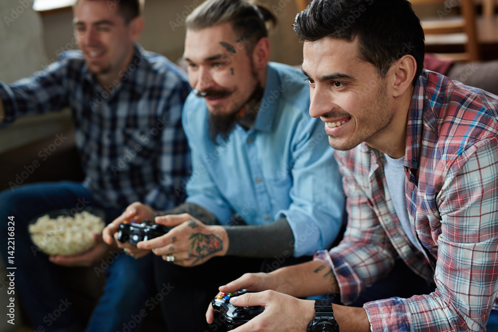 Portrait of three excited adult men enjoying video game competition and smiling cheerfully holding wireless controllers while sitting on couch in living room with popcorn