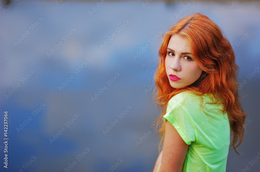 Portrait of a very beautiful serious brown eyed girls with bright red hair in a light green dress on a background of lake, close-up
