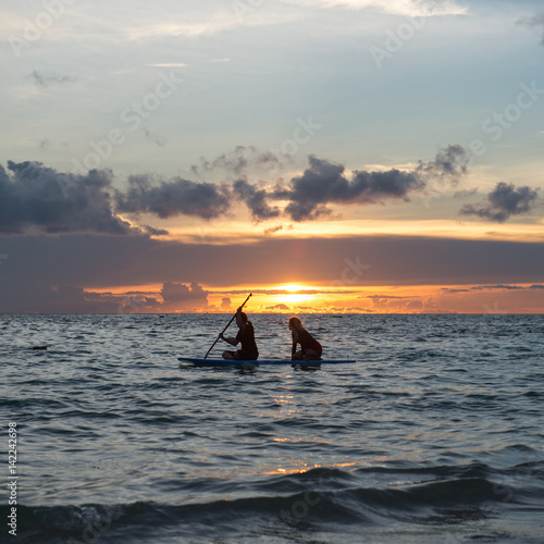 Two girls sits on paddle boarding on quiet sea at sunset
