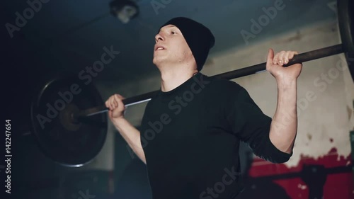 Muscular man doing crossfit training in a dark shadowy gym lifting weights holding a barbell at waist level in a health and fitness concept, movement round close shot. photo