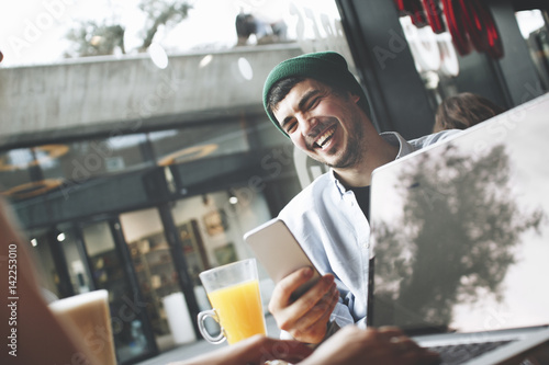 Joyful young man with a smartphone in his hand sits with his girlfriend who works for a laptop in a cafe next to a large window at lunchtime photo