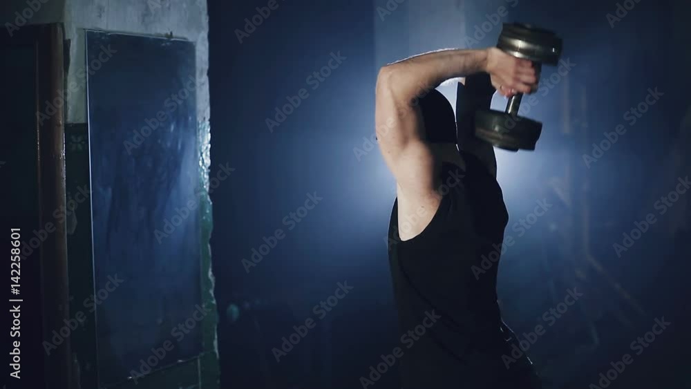 A man at a mirror raises a dumbbell. A dark gym with a blue backlight. A man at a mirror raises a dumbbell. A dark gym with a blue backlight.