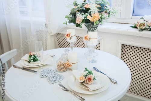 Interior design of white evening room with beautiful chandelier under served table with flowers on it. Big windows at the background. Vertical
