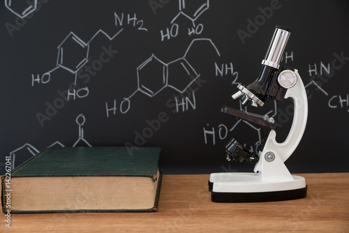 microscope and book on wooden table with science formula on black background