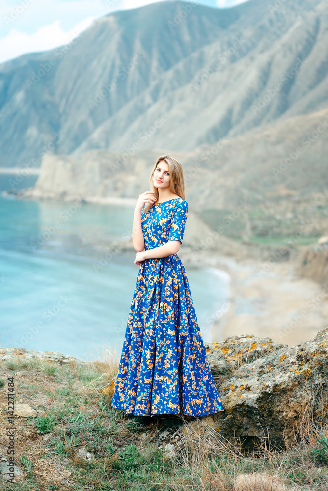 Beautiful Attractive Blond Caucasian Girl Model Stands Mountains and Sea Background in Long Blue Dress with Gold Yellow Pattern