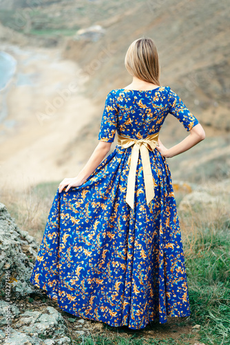 Beautiful Attractive Blond Caucasian Girl Model Stands Mountains and Sea Background in Long Blue Dress with Gold Yellow Pattern