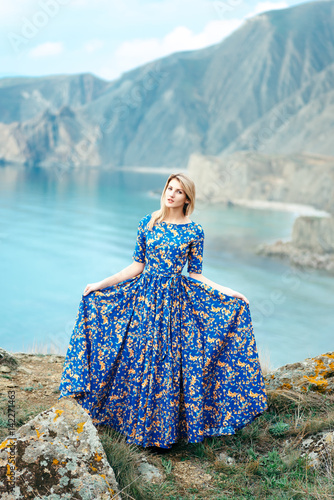 Beautiful Attractive Blond Caucasian Girl Model Stands Mountains and Sea Background in Long Blue Dress with Gold Yellow Pattern
