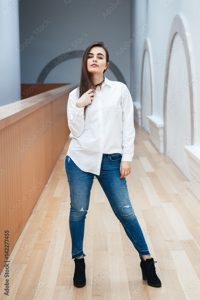 Portrait of sexy white Caucasian brunette young beautiful girl woman model  with long dark hair in white shirt and blue jeans indoor in hall at college  university standing full figure Stock Photo