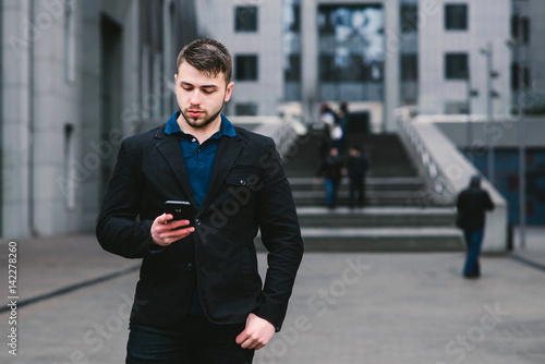 Young handsome businessman in a suit uses a smartphone on the background of modern urban landscape.