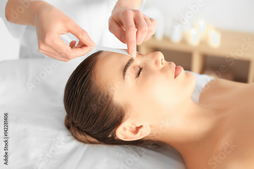 Beautician waxing young woman's eyebrows in spa center