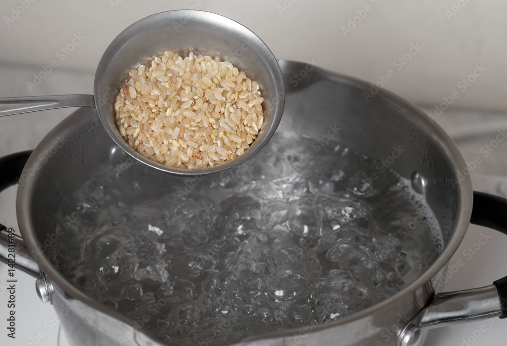 Raw brown rice in measuring cup over saucepan with boiling water