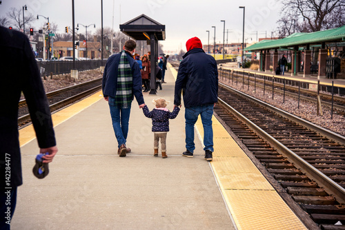 Back view of small girl holding hands with two young men on train platform