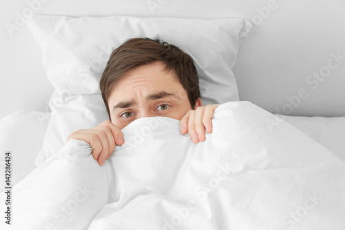 Young man hiding under blanket while lying in bed at home