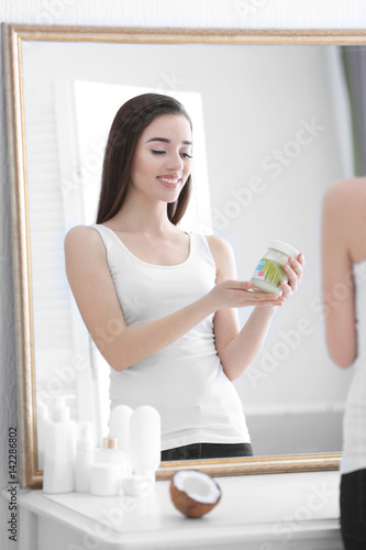 Beautiful young woman with jar of coconut oil standing in front of mirror at home