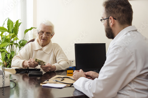 Lekarz pobiera opłatę od staruszki w prywatnym gabinecie lekarskim. Prywatna praktyka lekarska. Stara babcia odlicza pieniądze w gabinecie lekarskim. 