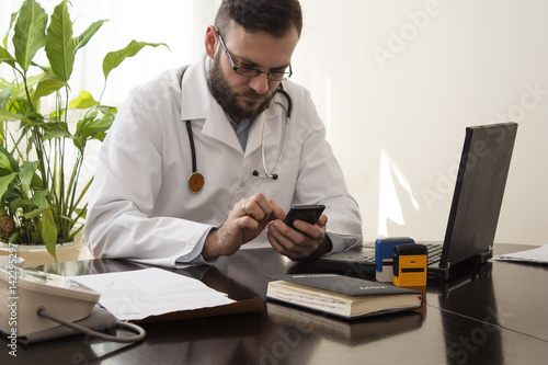 Lekarz w gabinecie lekarskim siedzi przy biurku z telefonem komórkowym w dłoni.Lekarz wybiera numer na telefonie.