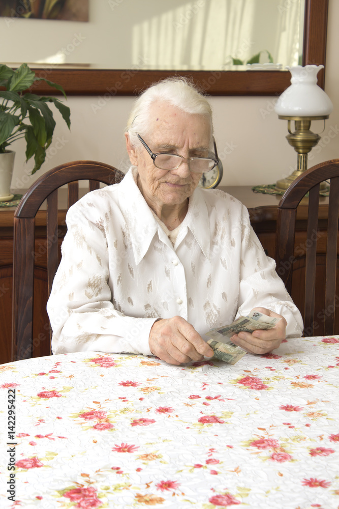 Stara kobieta ze smutną miną liczy pieniądze siedząc przy stole.Staruszka liczy pieniądze siedząc przy stole. - obrazy, fototapety, plakaty 