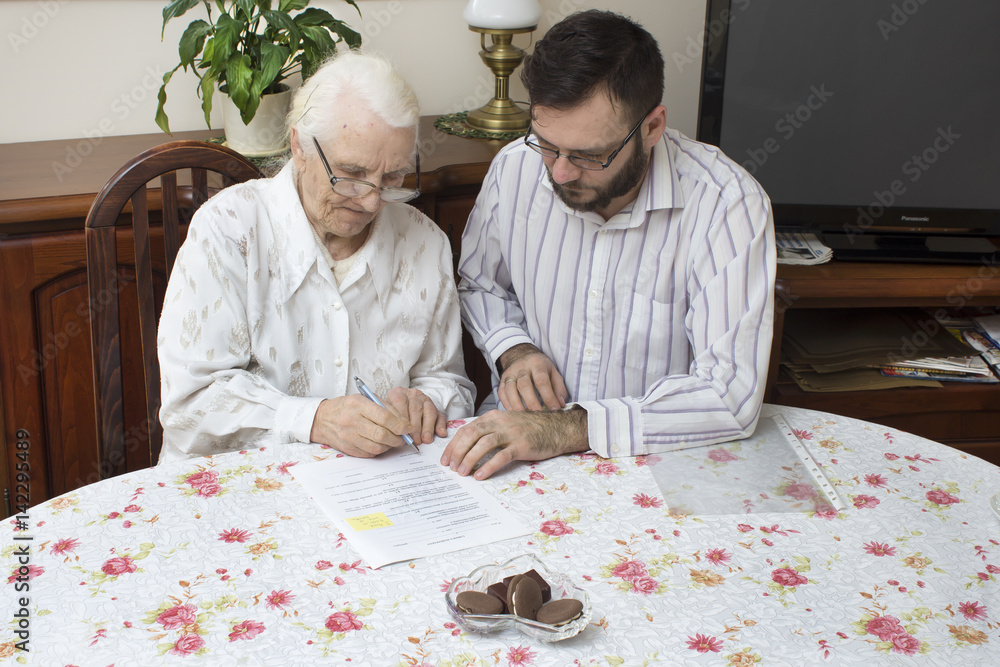 Umowa darowizny. Umowa kredytowa. Kredyt na starszą osobę. Staruszka podpisuje dokumenty siedząc przy stole. - obrazy, fototapety, plakaty 
