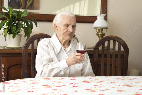 Babcia siedzi przy stole trzymając kieliszek z czerwonym winem. photo