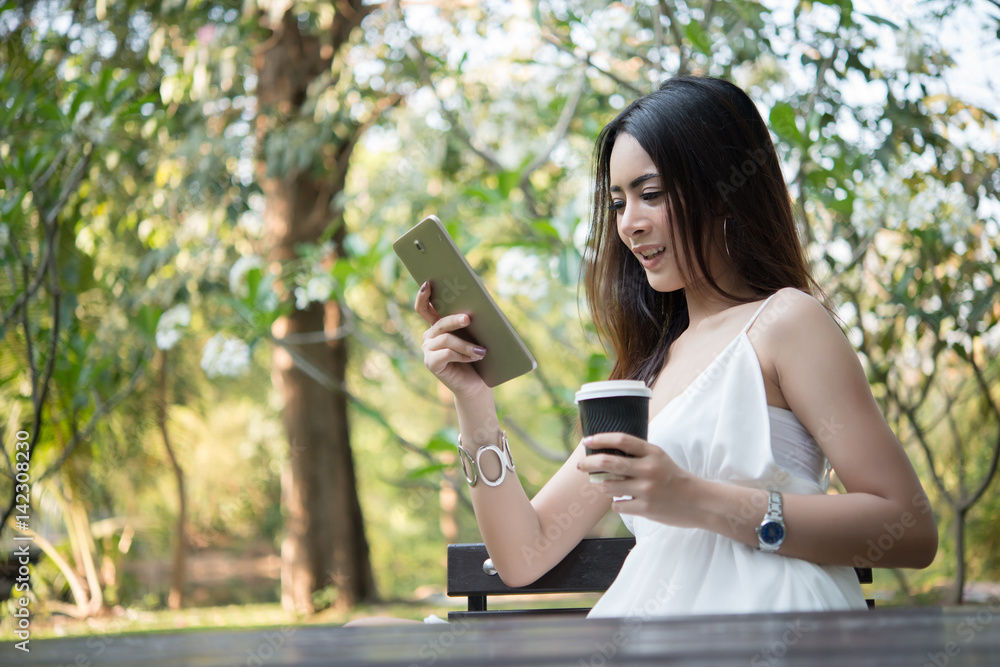 Young beautiful woman holding disposable coffee cup while text messaging through smartphone at nature outdoors. Young beautiful woman holding disposable coffee cup while text messaging through smartphone at nature outdoors.