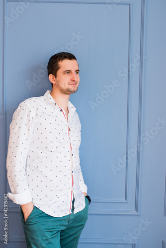 A handsome man in a white shirt with patterns and green pants leaned against the blue wall and looks out the window