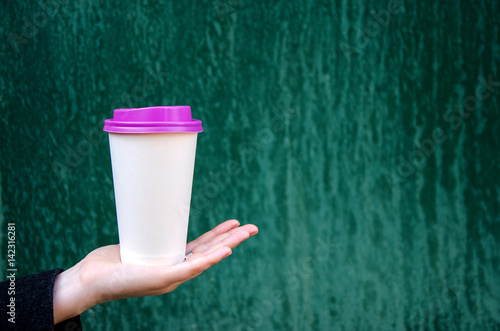 Fototapeta Naklejka Na Ścianę i Meble -  Mock up paper cup of coffee for branding .Cup of coffee with violet cap in woman's hand against green background. Good morning concept. Empty space for inscription.