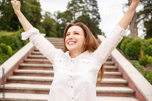 Happy woman with raised hands standing near stairs in park. Hispanic girl looking excited