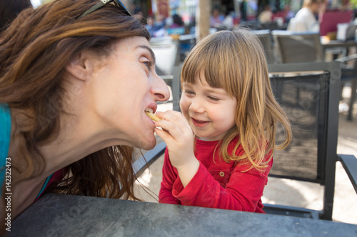 blonde three years old child face  with red shirt  feeding mother with cheese puff  laughing  sitting in terrace exterior bar cafe with grey table in park   