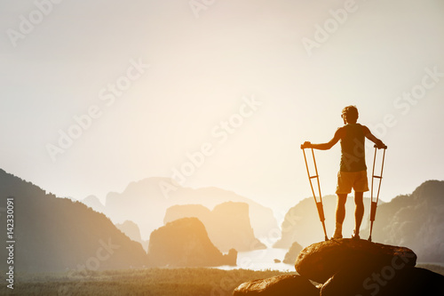 Fototapet Disabled man with crutches on big rock stands like winner