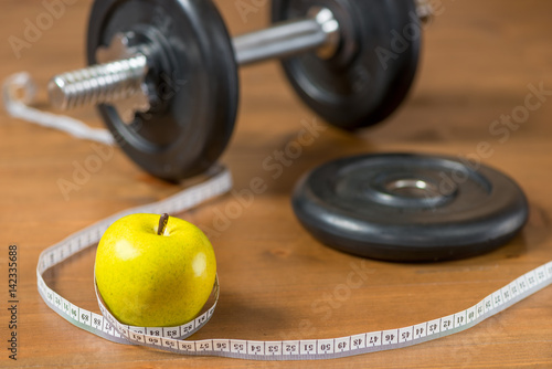 A green apple is wrapped in a centimeter against the background of a heavy dumbbell on a wooden floor